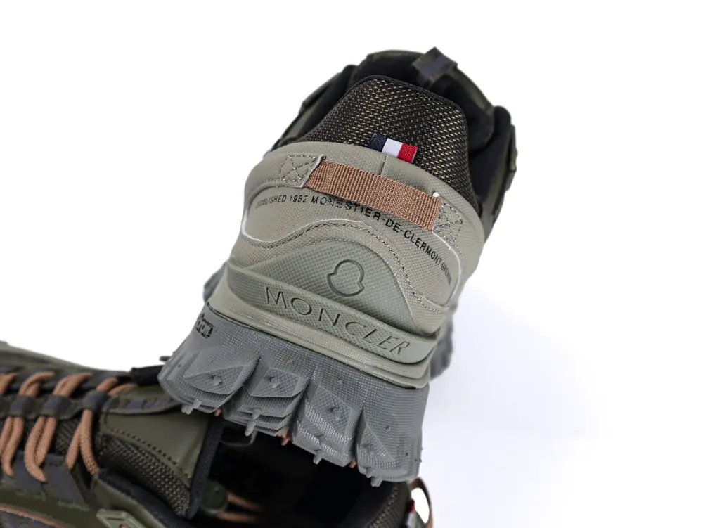 Close-up of a Moncler shoe, featuring green and brown materials, textured sole, and a small tri-color fabric tab.