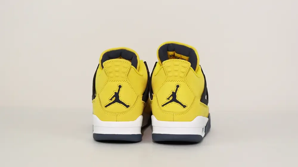 A rear view of both shoes of the Jordan 4 Retro ‘Lightning’ (2021), showcasing the overall yellow leather upper and the metallic black Jordan logo on the heel.